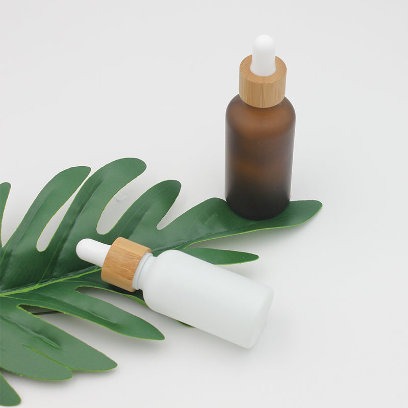 Frosted glass dropper bottles in amber and white, featuring bamboo caps, placed on a green leaf.
